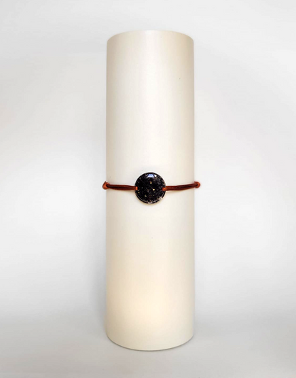 Image d'un modèle de bracelet Mimi avec son cordon satiné de couleur cuivrée et sa pastille ronde en résine incrustée de fleurs naturelles séchées noires et de paillettes de feuille de cuivre dorée.