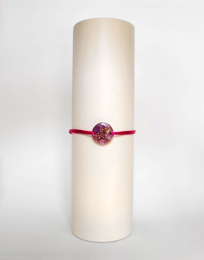 Image d'un modèle de bracelet Mimi avec son cordon satiné de couleur fushia et sa pastille ronde en résine incrustée de poudre de mica rose et de paillettes de feuille de cuivre dorée.