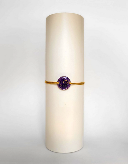 Image d'un modèle de bracelet Mimi avec son cordon satiné de couleur jaune et sa pastille ronde en résine incrustée de fleurs naturelles séchées violettes et de paillettes de feuille de cuivre dorée.