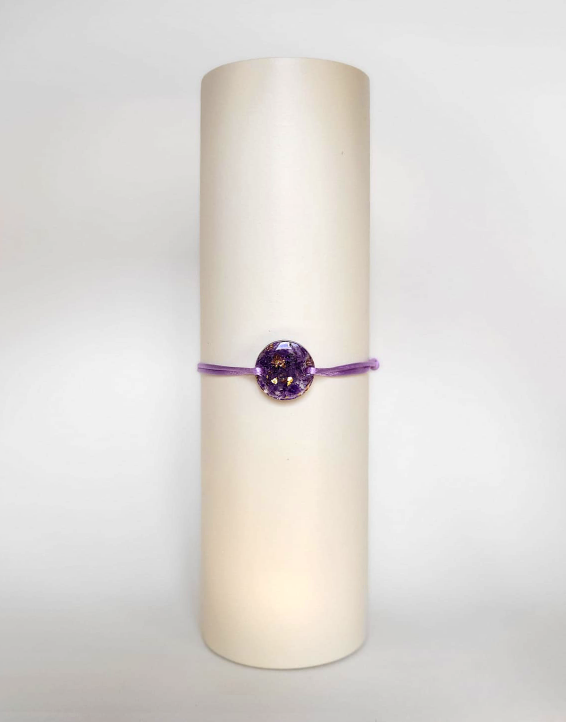 Image d'un modèle de bracelet Mimi avec son cordon satiné de couleur lilas et sa pastille ronde en résine incrustée de fleurs naturelles séchées violettes et de paillettes de feuille de cuivre dorée.