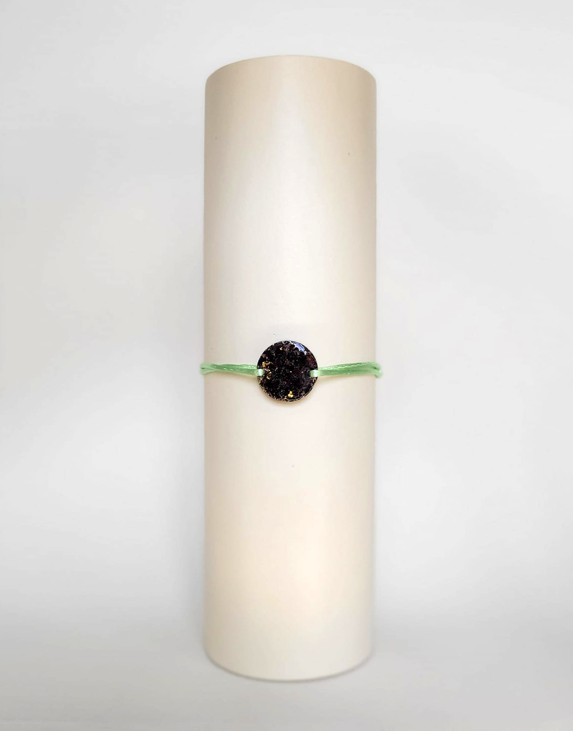 Image d'un modèle de bracelet Mimi avec son cordon satiné de couleur verte pomme et sa pastille ronde en résine incrustée de fleurs naturelles séchées noires et de paillettes de feuille de cuivre dorée.