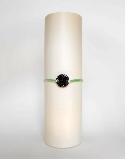 Image d'un modèle de bracelet Mimi avec son cordon satiné de couleur verte pomme et sa pastille ronde en résine incrustée de fleurs naturelles séchées noires et de paillettes de feuille de cuivre dorée.
