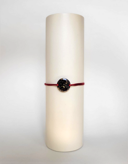 Image d'un modèle de bracelet Mimi avec son cordon satiné de couleur rouge et sa pastille ronde en résine incrustée de fleurs naturelles séchées noires et de paillettes de feuille de cuivre dorée.