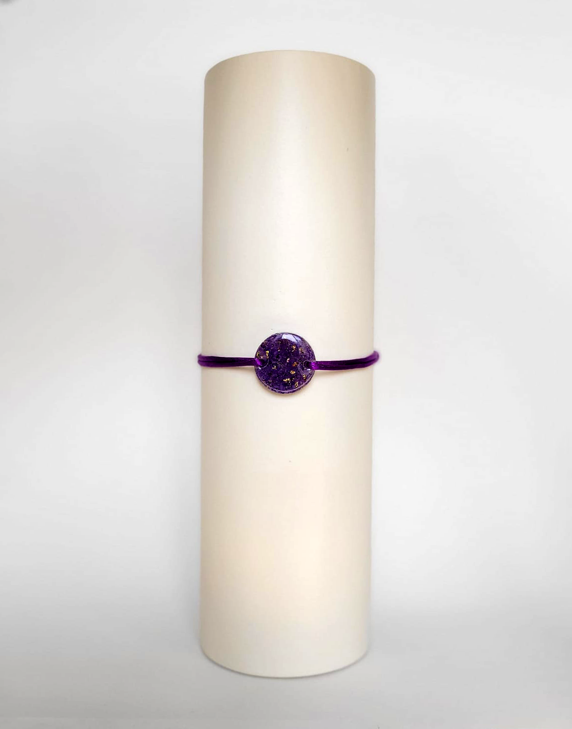 Image d'un modèle de bracelet Mimi avec son cordon satiné de couleur violette et sa pastille ronde en résine incrustée de fleurs naturelles séchées violettes et de paillettes de feuille de cuivre dorée.