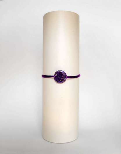 Image d'un modèle de bracelet Mimi avec son cordon satiné de couleur violette et sa pastille ronde en résine incrustée de fleurs naturelles séchées violettes et de paillettes de feuille de cuivre dorée.